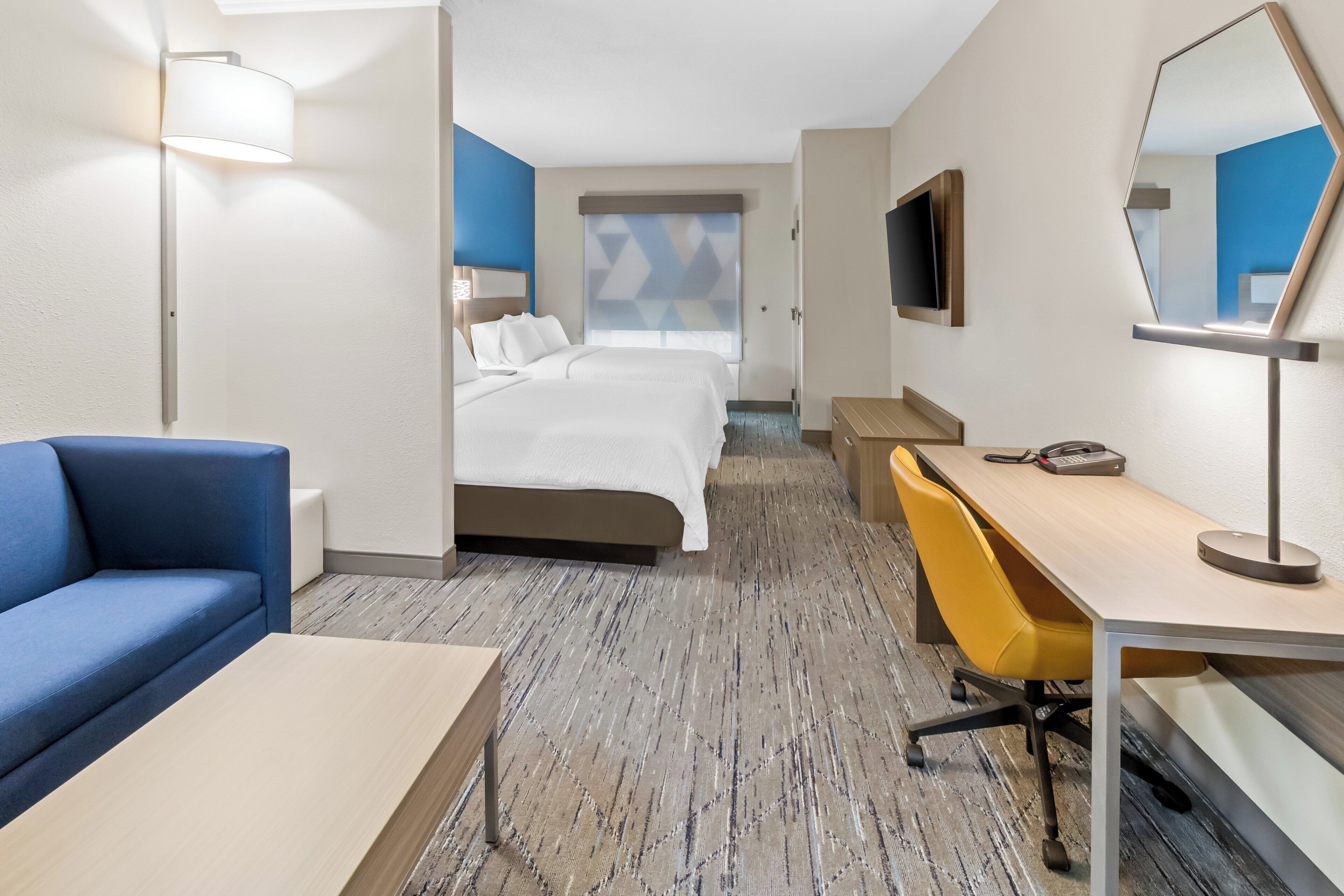 Ein Hotelzimmer mit Bett, Schreibtisch, Stuhl, Sofa, Fernseher und Spiegel.