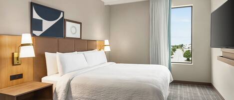 A hotel room with a large bed, two bedside lamps, a wooden nightstand, a flat-screen TV, and a window with a view of the outside.