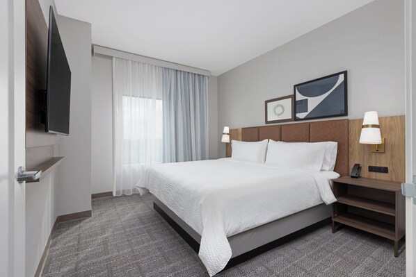 Habitación de hotel con cama, televisor, mesita de noche con lámpara y ventana con cortinas.