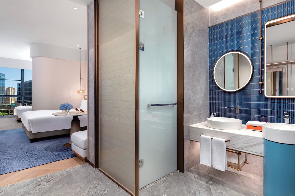 A modern hotel room with a large bed, a separate bathroom with a blue tiled wall, and a city view.