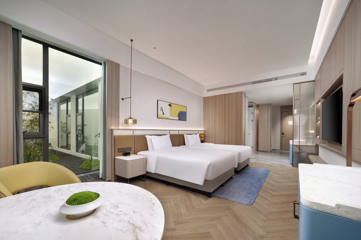 A modern hotel room with a large bed, bedside tables, a flat-screen TV, and a large window offering a view of the outdoors.