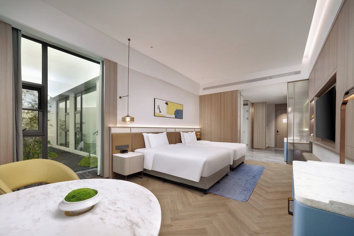 A modern hotel room with a large bed, bedside tables, a flat-screen TV, and a large window offering a view of the outdoors.