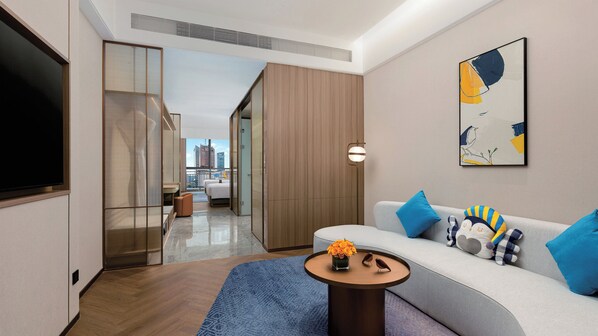 A modern hotel room with a sofa, a coffee table, a TV, and a view of the cityscape.