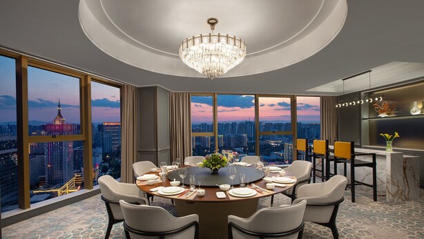 A modern dining room with a round table, chairs, and a chandelier. It features large windows with a city view.