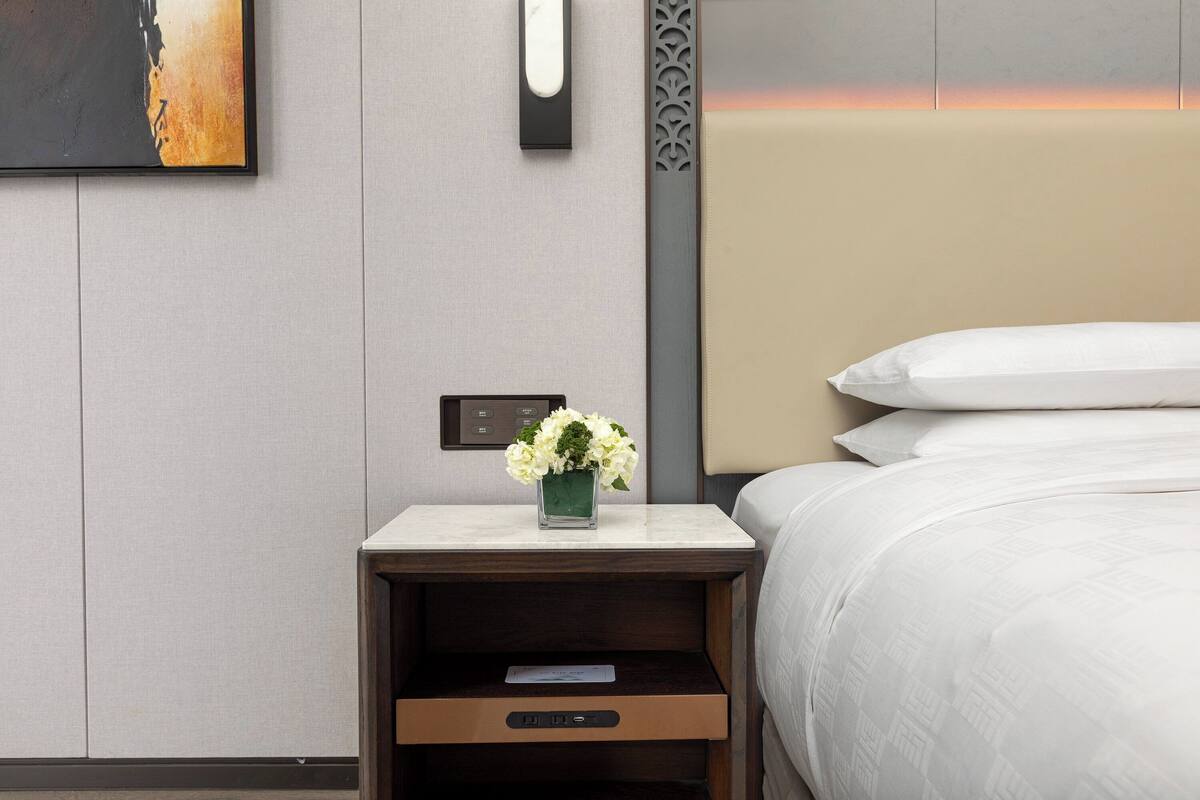 Ein ordentlich eingerichtetes Hotelzimmer mit einem Bett, einem Nachttisch mit Blumenvase, einer Wanduhr und einer Bedienkonsole.
