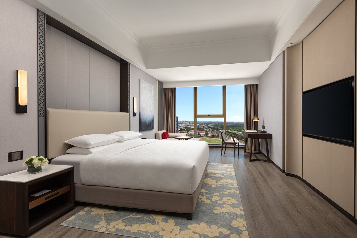 A modern hotel room with a large bed, a desk, a TV, and a view of the city.