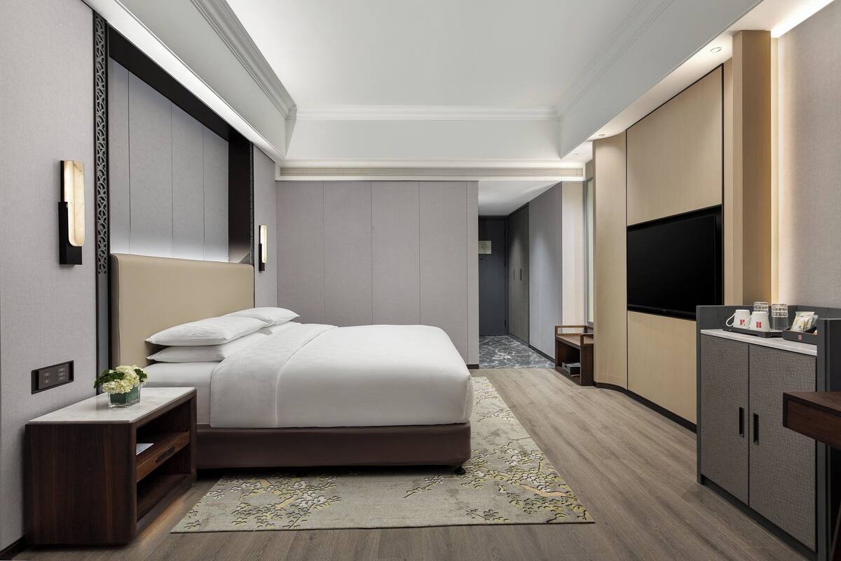 Ein modernes Hotelzimmer mit einem großen Bett, einem Fernseher, einer Minibar und einem kleinen Schreibtisch mit Pflanze.
