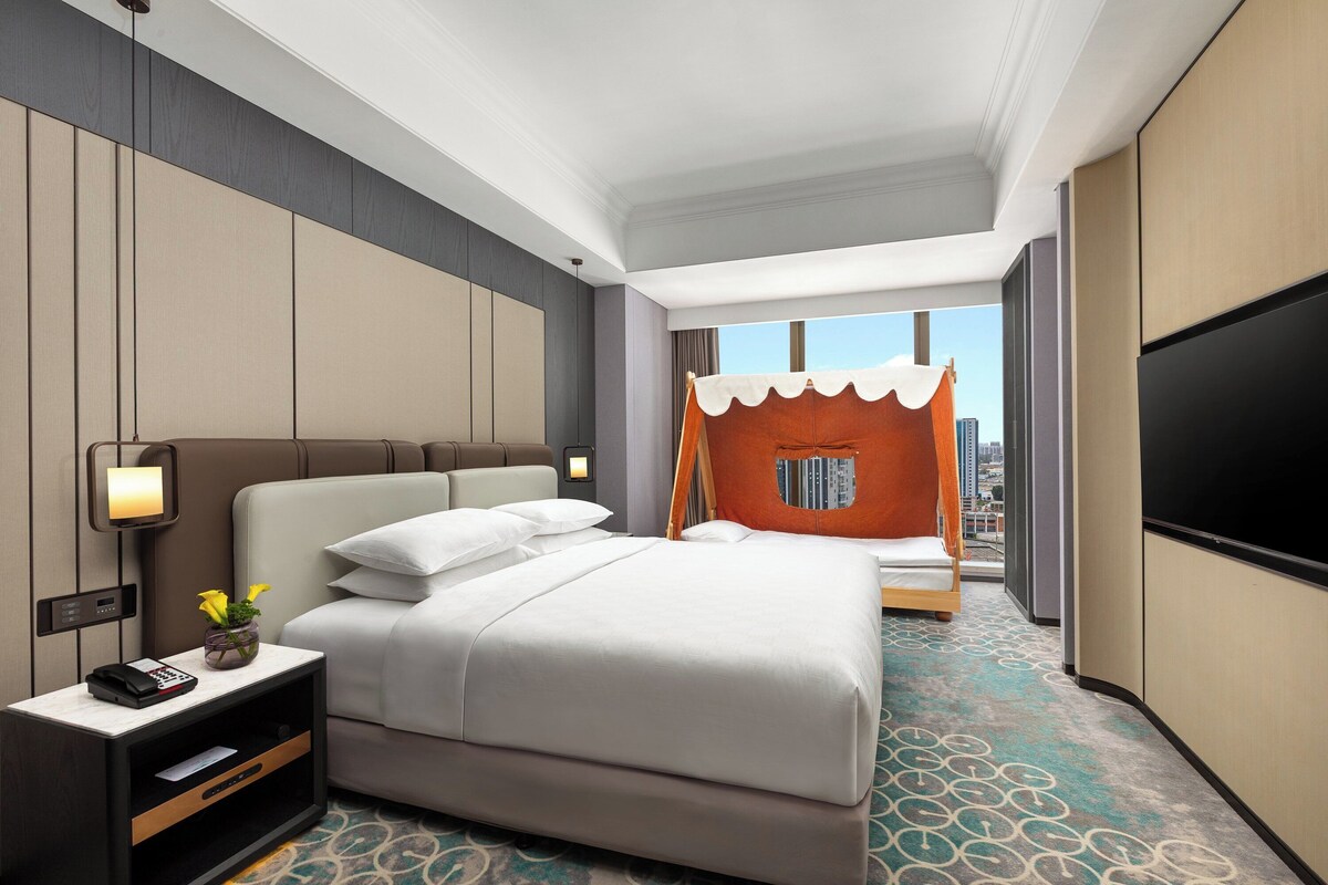 A hotel room with a large bed, a TV, a bedside table with a lamp, and a view of the city.