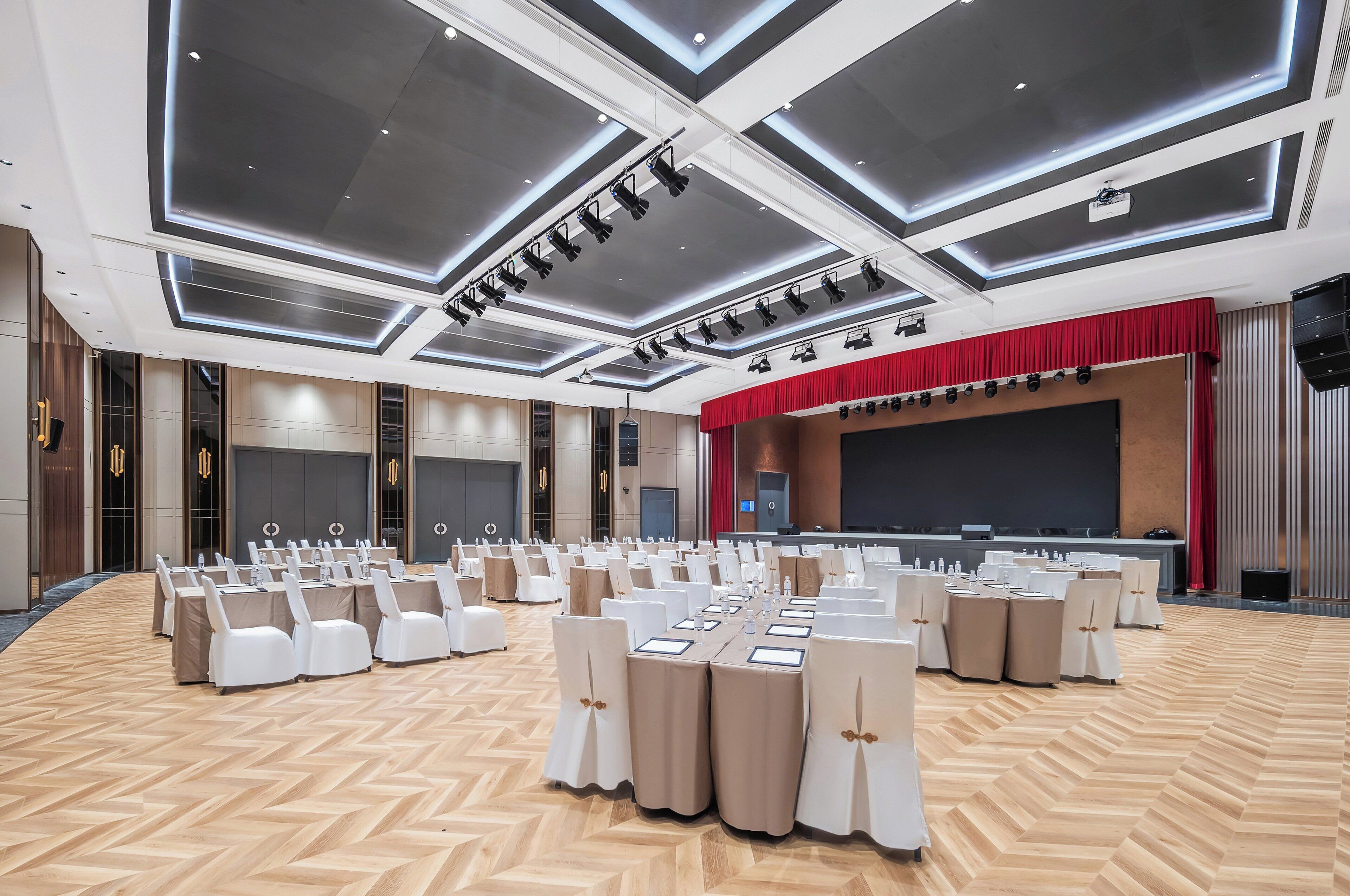 A conference room with rows of tables and chairs, a stage with a red curtain, and a large screen.