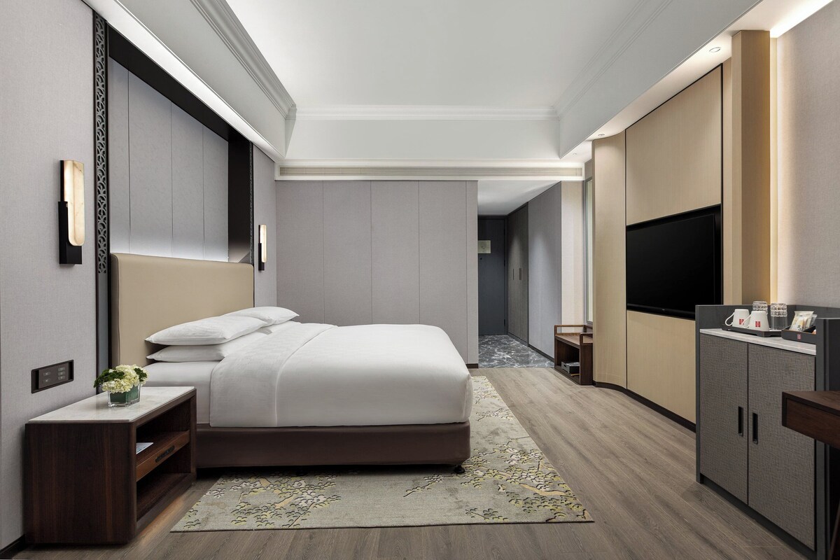 A modern hotel room with a large bed, a TV, a minibar, and a small desk with a plant.