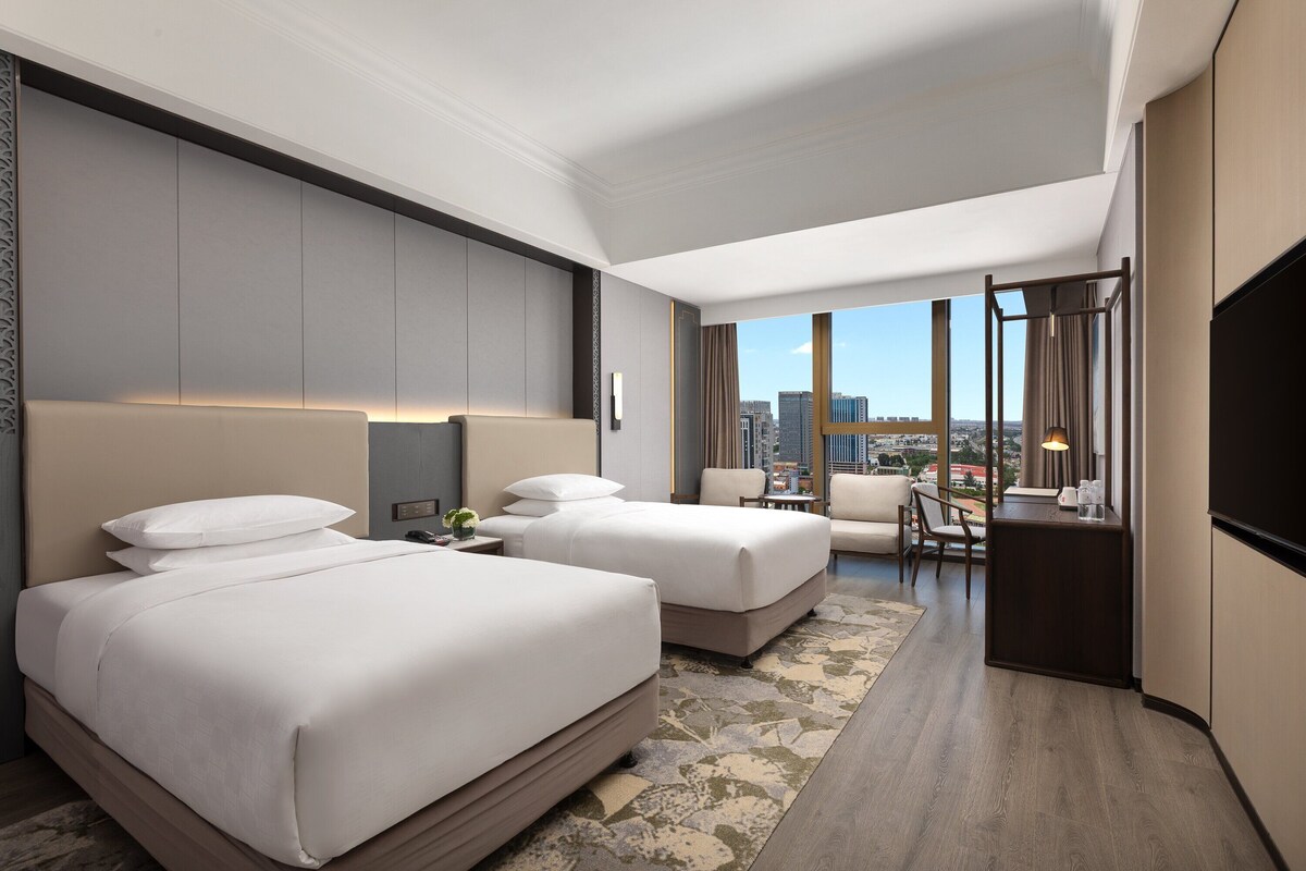 A hotel room with two beds, a desk, and a view of the city.