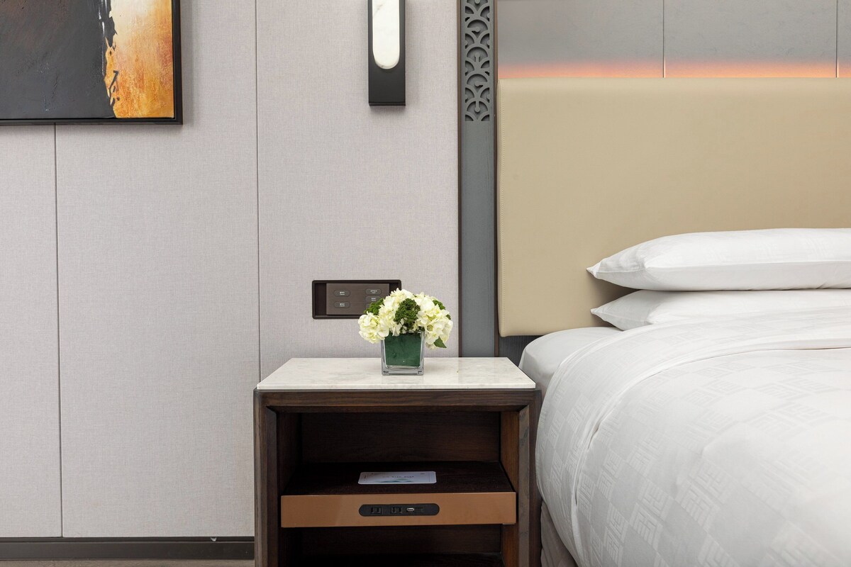 A neatly made hotel room with a bed, a nightstand with a vase of flowers, a wall clock, and a control panel.
