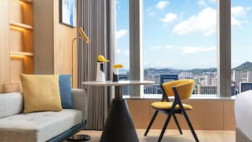 A modern hotel room with a sofa, a dining table, and a chair. The room has a large window with a city view.