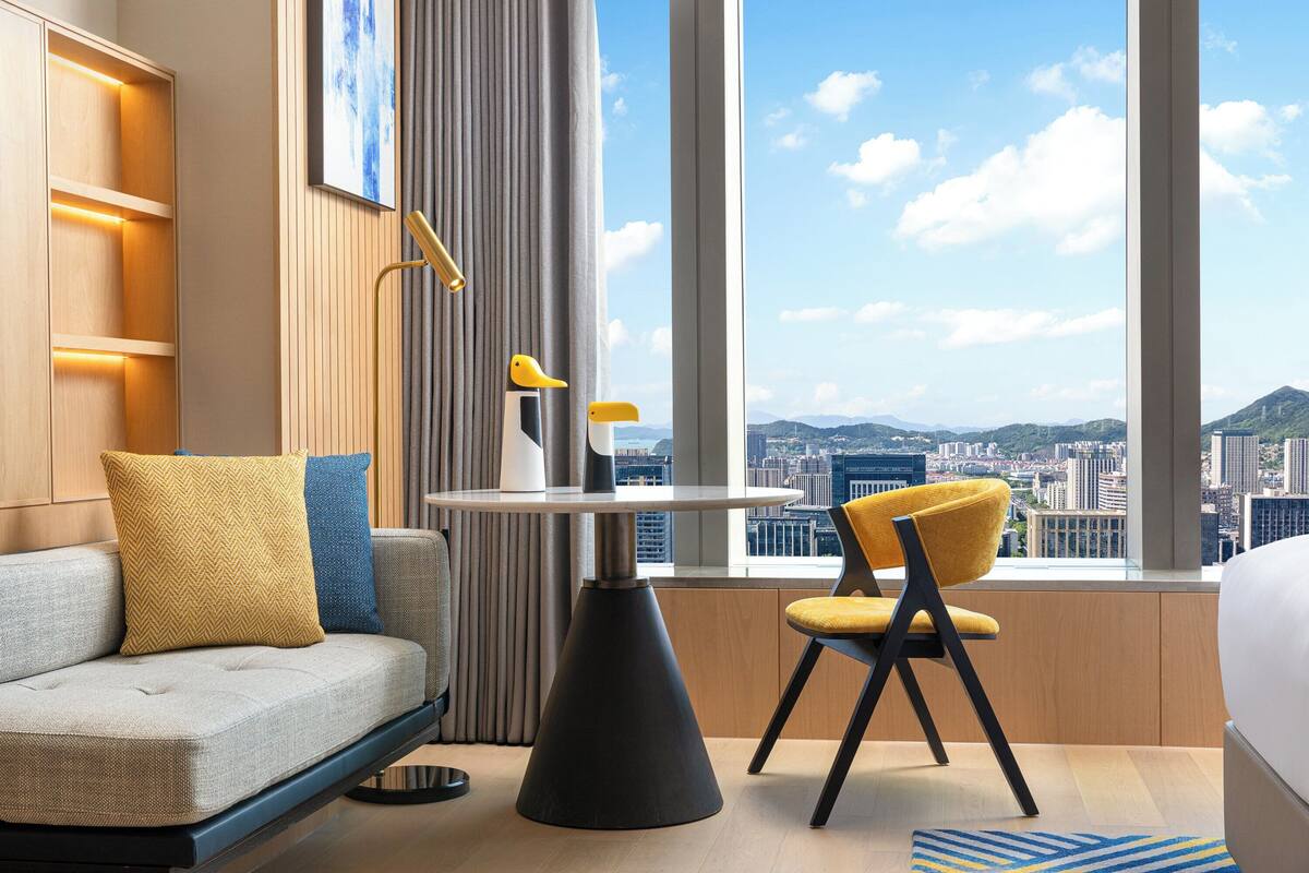 Habitación de hotel moderna con sofá, una mesa de comedor y una silla. La habitación tiene un ventanal con vistas a la ciudad.