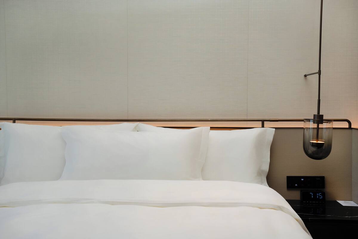 Una cama bien hecha con sábanas blancas, una mesita de noche negra con un reloj digital y una lámpara colgante.