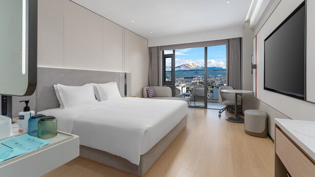 Habitación de hotel moderna con una cama grande, un escritorio, un televisor y un balcón con vistas a la montaña.