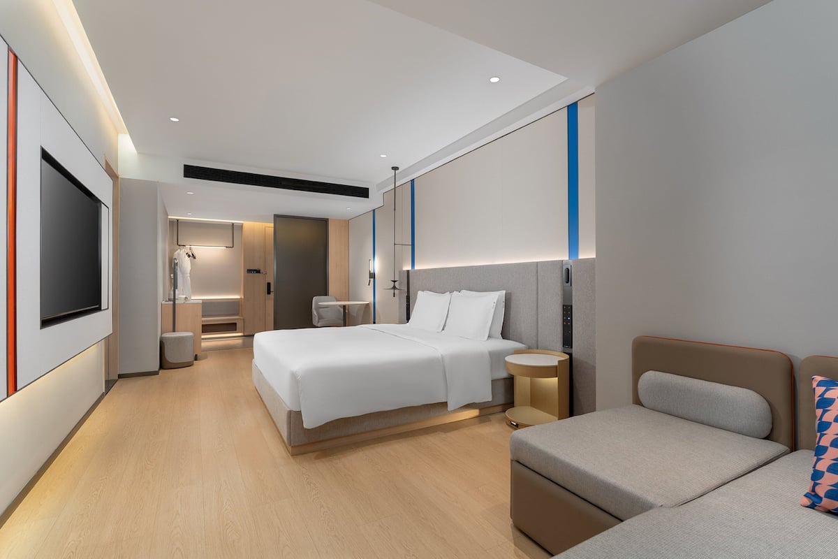 Ett modernt hotellrum med en stor säng, en mindre soffa, ett sängbord, en TV och ett badrum med dusch och toalett.