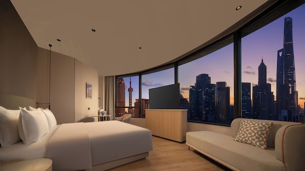 Ein modernes Hotelzimmer mit einem großen Bett, einer Couch, einem Schreibtisch und Blick auf eine Stadtsilhouette.