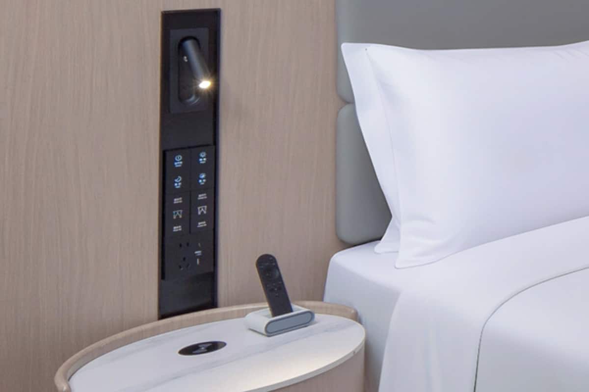 A hotel room with a control panel for room amenities, a bedside table with a remote control, and a neatly made bed.