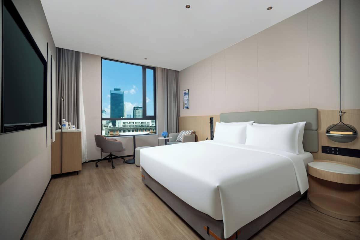 A modern hotel room with a large bed, a desk, a chair, and a view of the cityscape through a large window.