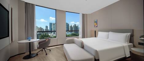 A modern hotel room with a large bed, a small desk, a chair, and a view of the cityscape through large windows.
