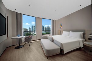 Una habitación de hotel moderna con una cama grande, un escritorio pequeño, una silla y vistas a la ciudad a través de amplios ventanales.