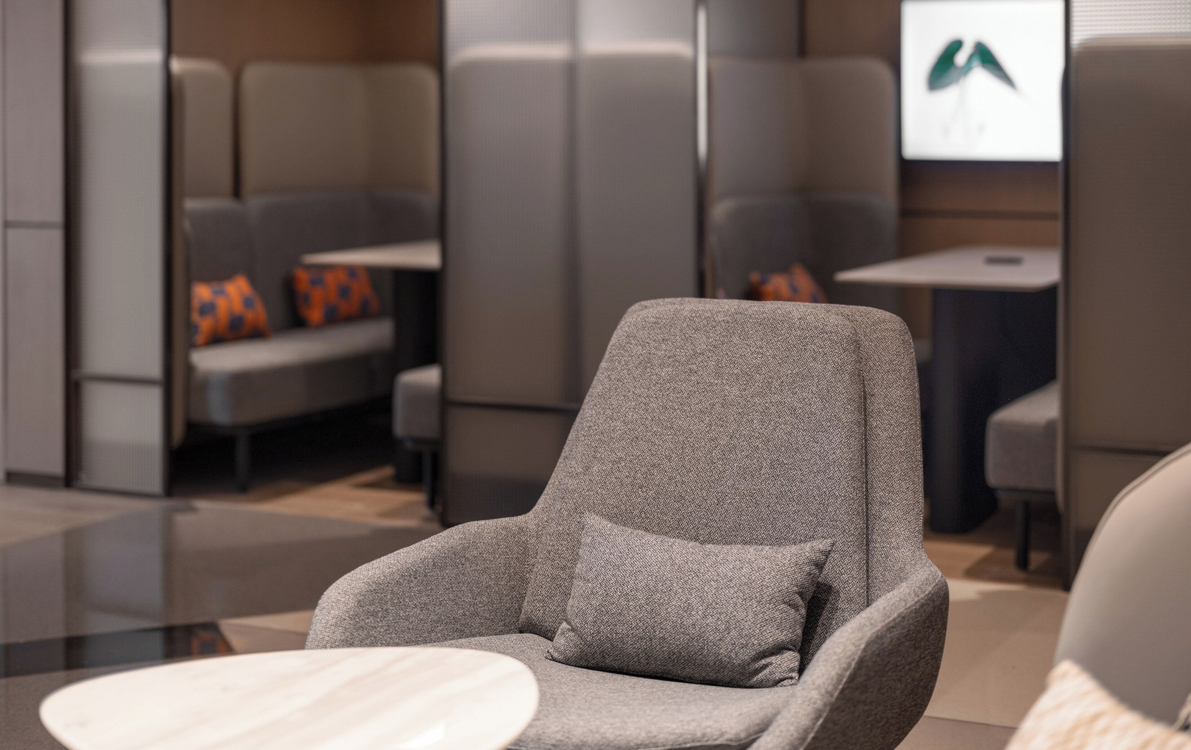 A modern lounge area with a grey armchair and a small table, a seating area with orange cushions, and a wall-mounted screen displaying a logo.