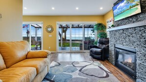 A living room with a leather sofa, a fireplace, a large TV, and a view of the outdoors through glass doors. - Peaceful Point Lakehouse with Hot tub (Leamington)