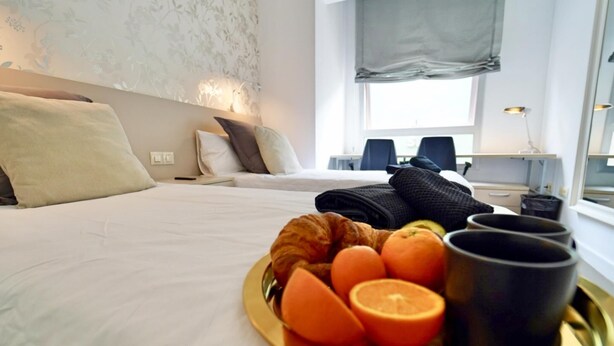 Habitación de hotel con una cama, una bandeja con cruasanes, naranjas y una taza, además de un escritorio con lámpara y silla.