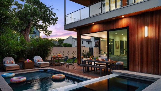 A modern outdoor pool area with lounge chairs and a dining table, surrounded by wooden paneling and a large tree. - Casa Sol - Heated Pool, Outdoor Living & Design (Austin)