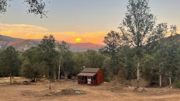 Une petite cabane en bois avec un porche, entourée d’arbres et avec des collines en arrière-plan.