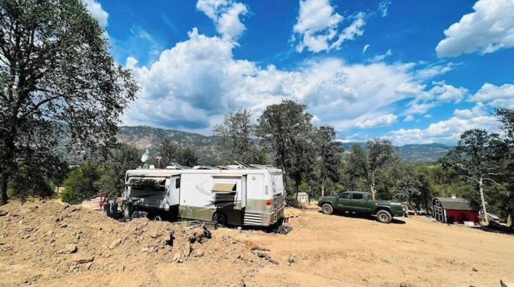A white RV parked on a dirt road with a green truck nearby, surrounded by trees and hills in the background.