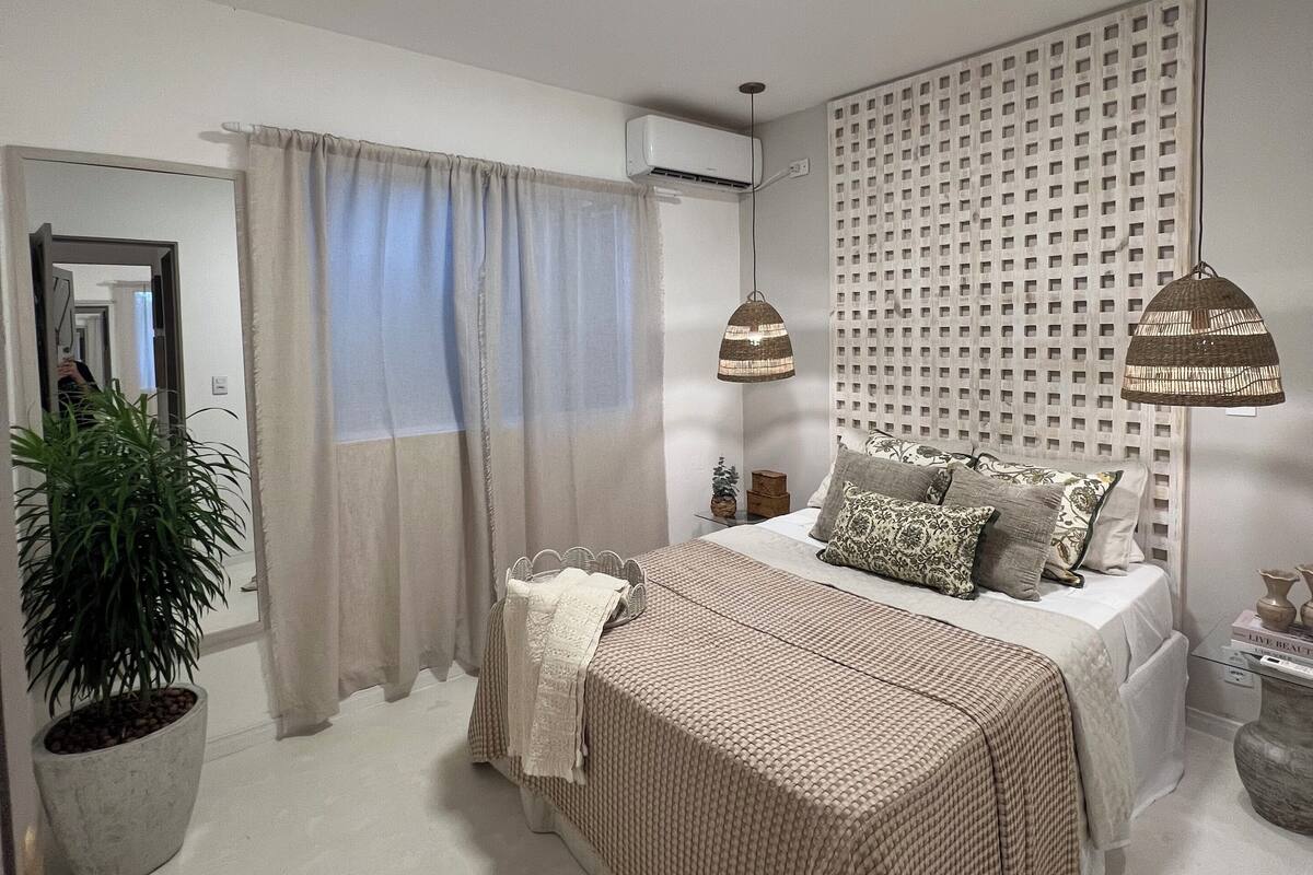 Ein Schlafzimmer mit Bett, Nachttischen, einer Topfpflanze, einem Fenster mit Vorhängen, einer Klimaanlage und einer Wandleuchte.