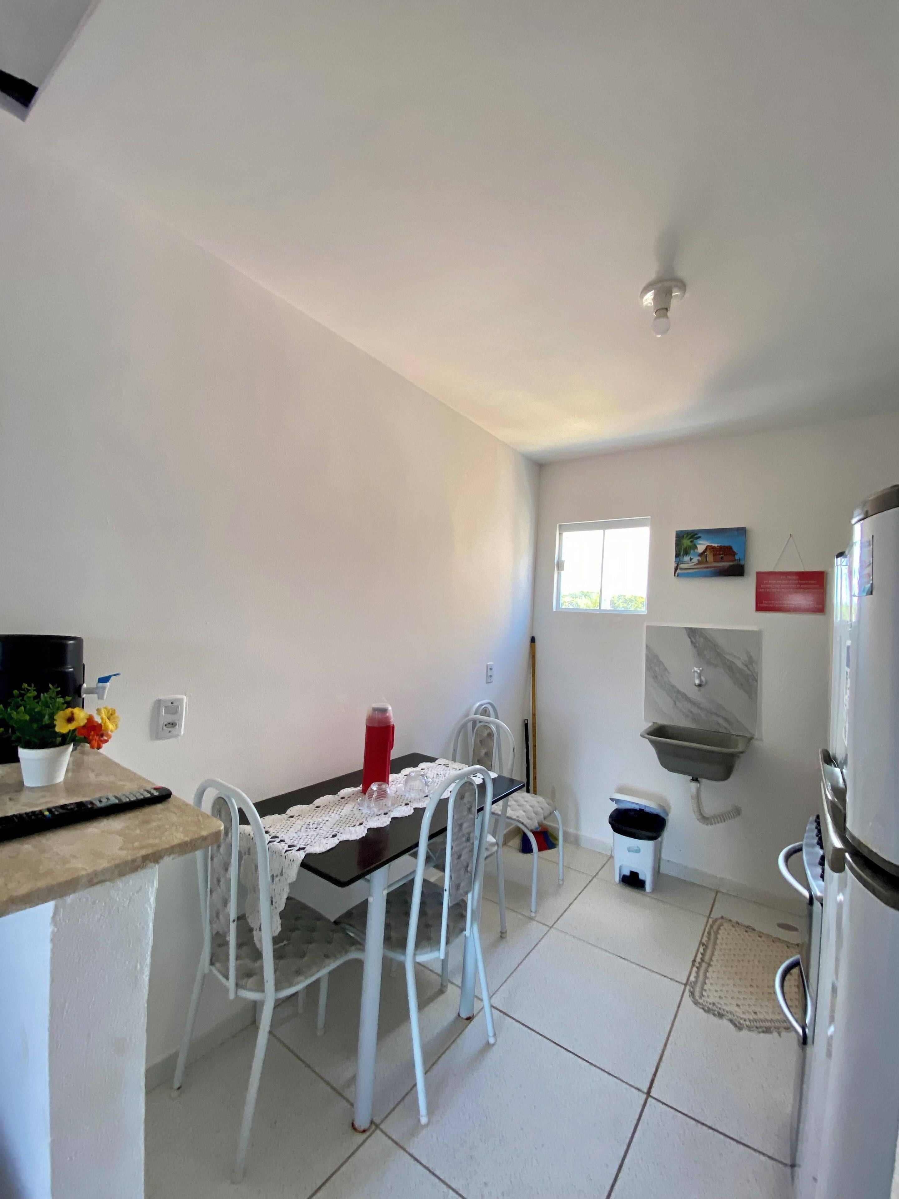A small, clean kitchen with white walls, a tiled floor, a table with chairs, a refrigerator, a sink, and a window.