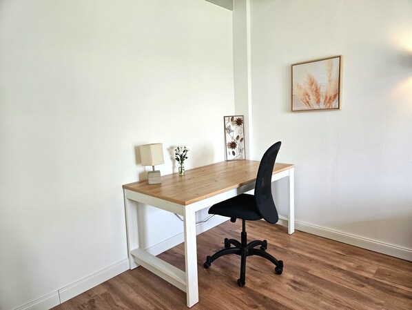 A small, white-framed desk with a wooden top, a black office chair, a small lamp, and a decorative metal piece on the wall.