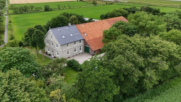 A large, two-story house with a red-tiled roof, surrounded by lush greenery and fields.
