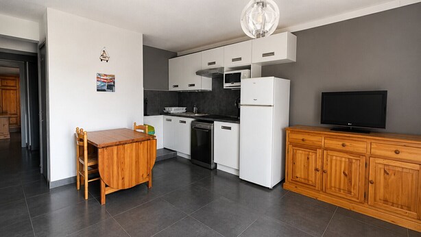 A modern kitchen with white cabinetry, a wooden dining table, a black refrigerator, and a television on a wooden cabinet.