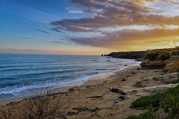 A coastal landscape with a sandy beach, rocky outcrops, and a clear sky.