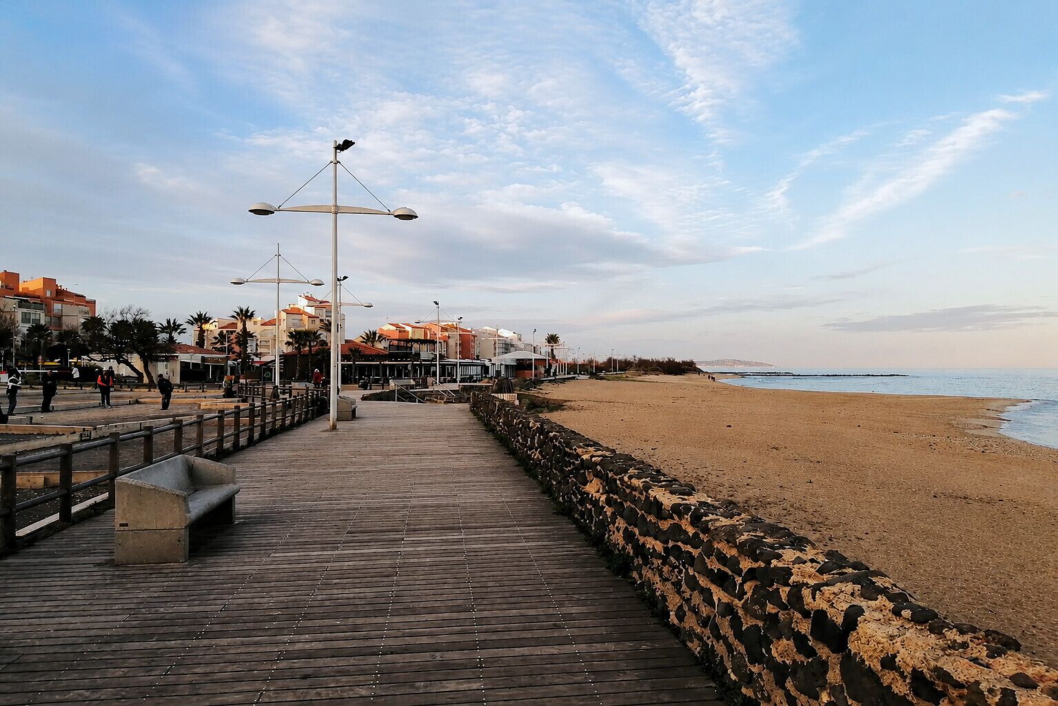 A wooden boardwalk leading to a sandy beach with a stone wall on one side and buildings in the background.