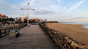 A wooden boardwalk leading to a sandy beach with a stone wall on one side and buildings in the background.