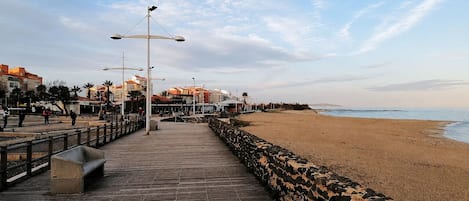 Een houten looppad dat naar een zandstrand leidt, met een stenen muur aan één kant en gebouwen op de achtergrond.