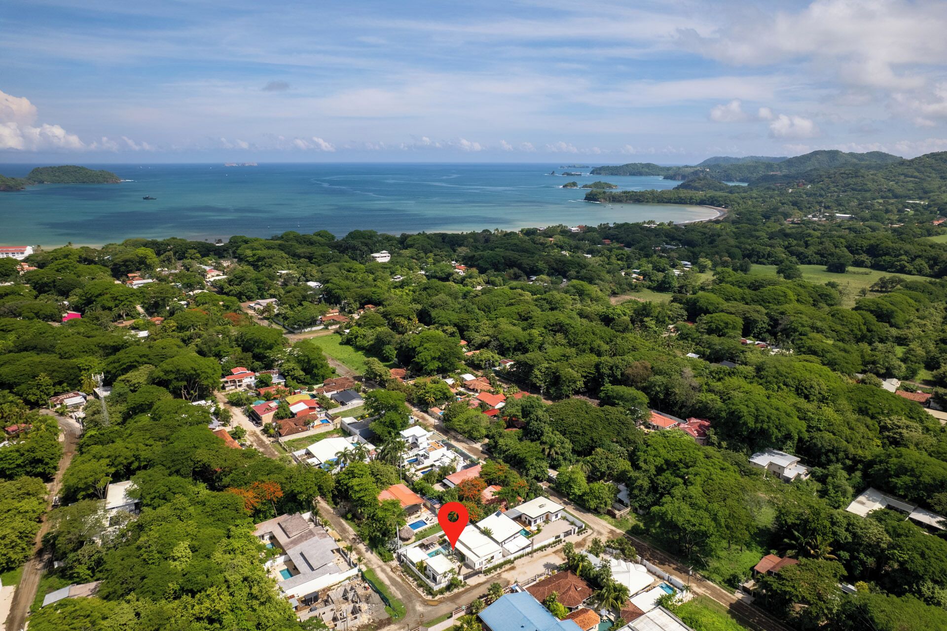 An aerial view of a coastal town with dense greenery, a mix of residential and commercial buildings, and a clear view of the ocean.