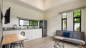 Une cuisine moderne équipée d’un réfrigérateur encastré, d’une machine à café et d’une fenêtre donnant sur un espace verdoyant. On y trouve un coin repas avec une table et des chaises en bois, ainsi qu’un canapé gris agrémenté d’un coussin bleu.