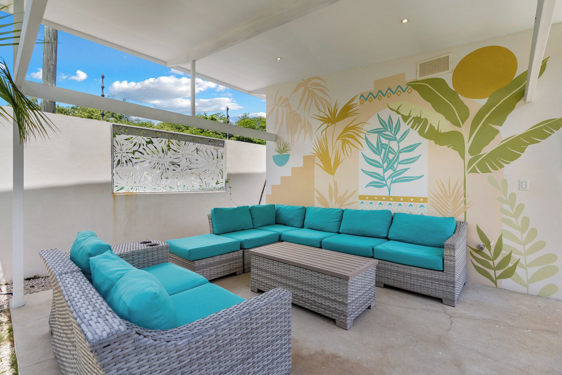 Un espace extérieur moderne, aménagé avec un canapé d’angle et deux fauteuils, un mur décoratif orné de motifs de feuilles tropicales et une structure de plafond blanche.