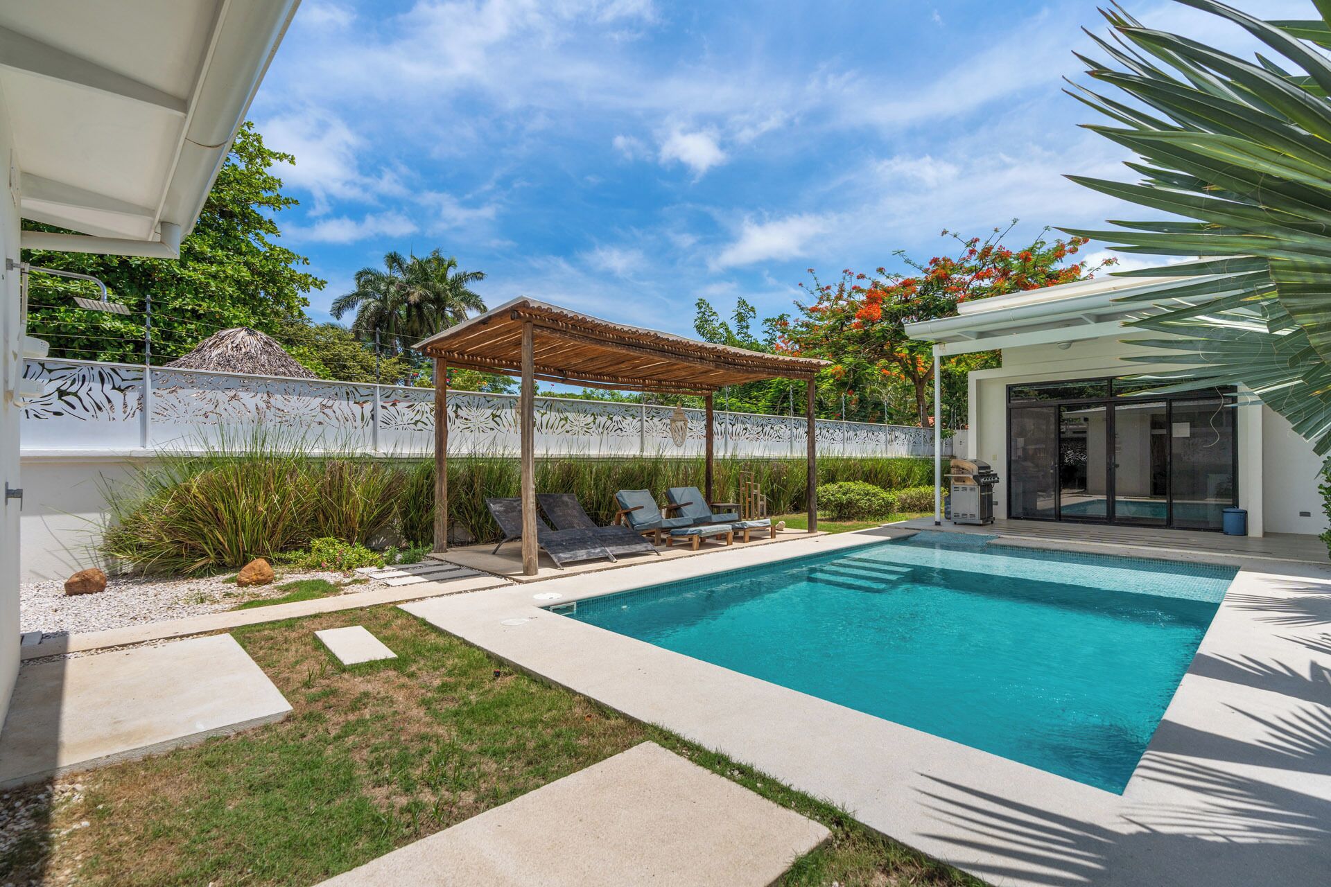 Un área de piscina moderna al aire libre con una pérgola, sillas de descanso y un porche cubierto.