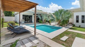 Un espace piscine extérieur moderne, agrémenté d’une pergola en bois, de chaises longues et d’un jardin bien entretenu avec des palmiers.