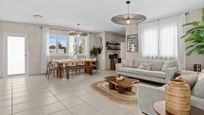 A spacious living area with a grey sofa, wooden coffee table, and a dining table with chairs. There are two pendant lights, a large plant, and a bookshelf. - 2 Boho Apartments in 1 Listing: With Pool and Steps to Beach (San Juan)