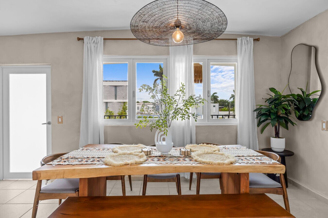 A dining area with a wooden table, white curtains, a hanging light fixture, and a view of buildings and trees through large windows.