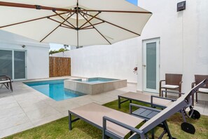 Un área de piscina moderna al aire libre con un jacuzzi, sillas de descanso y una sombrilla grande.