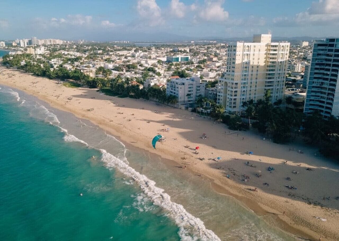 An aerial view of a beach with a parasail, buildings, and a coastline.